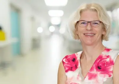 Dr. Sara Watkin, a woman with short, light hair and glasses, smiles while wearing a white dress adorned with pink and red floral patterns. She stands in a brightly lit hallway, the background softly out of focus.