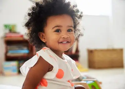 A smiling toddler with curly hair, wearing a white dress with orange polka dots, sits in a cozy room reminiscent of a Healthcare Clinic Cayman, complete with bookshelves and a wicker basket in the background.