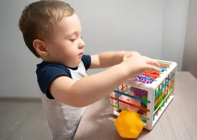 A toddler from their babies' first year places a toy into a white plastic cage-like container with multicolored slats, while other toys are scattered on a table.