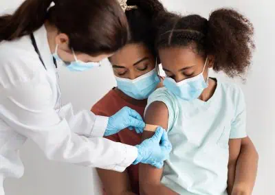 A healthcare worker in a white coat and gloves places a band-aid on the upper arm of a young girl wearing a light blue shirt, having just received her immunisations, as a woman watches closely. All are wearing face masks.