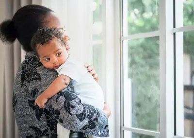 A person holding a baby looks out of the window, lost in thoughts about Watkin's out-of-hours service.