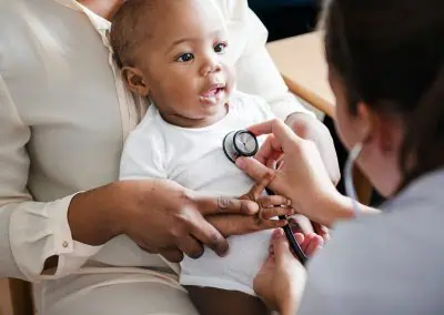 A baby, held by an adult, is being examined by a healthcare professional using a stethoscope.