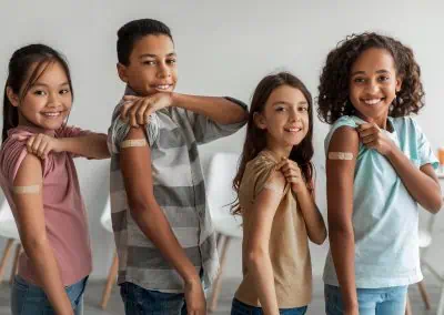 Four children standing in line indoors, showing bandages on their upper arms, indicating they have recently received immunisations.