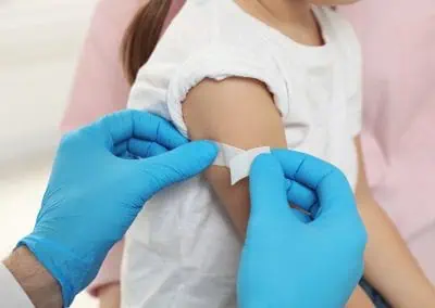 A child receiving a bandage on their upper arm following immunisations from a healthcare professional wearing blue gloves.