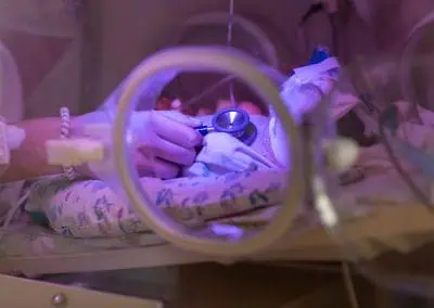 Medical professional using a stethoscope to examine a newborn in an incubator under purple lighting.