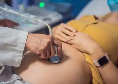 A medical professional performs an ultrasound on a pregnant woman's abdomen while she lies on an examination table, wearing a yellow shirt and a smartwatch.