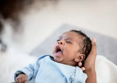 A newborn baby in a light blue outfit yawns while being gently cradled by a hand.