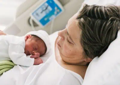 A person rests in a hospital bed with their newborn baby sleeping on their chest.