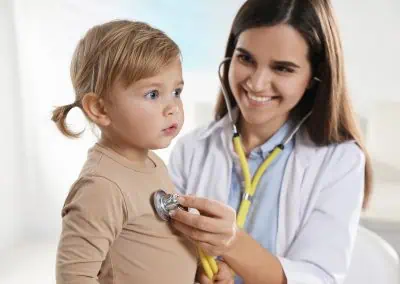 A paediatrician in Cayman uses a stethoscope to check a young child's heartbeat, both seated with the child looking to the side.