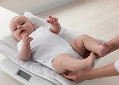 A baby in a white onesie lies on a padded scale while a caregiver adjusts the baby's legs.