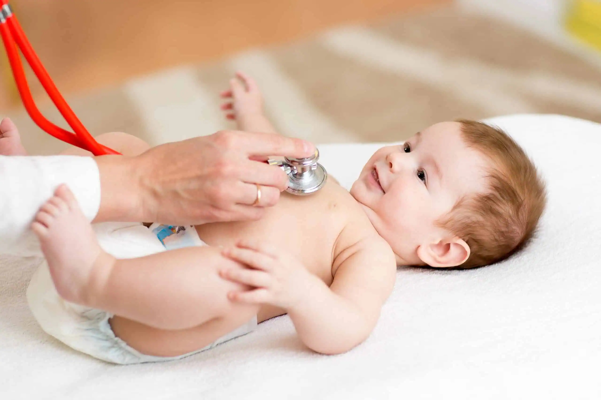 A baby lies on a white blanket while a person uses a stethoscope to examine the baby's chest.