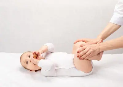 A baby in a white onesie lies on its back, holding a toy, while an adult's hands gently hold the baby's legs.