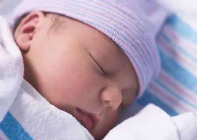 Newborn baby sleeping peacefully, wrapped in a white blanket, wearing a pink and blue striped hat.