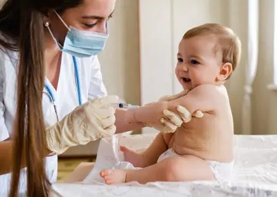 A healthcare professional wearing a mask and gloves administers immunisations to a baby sitting on a medical examination table.