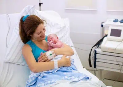 A woman rests in a hospital bed, cradling a newborn baby in her arms. Medical equipment is visible in the background.