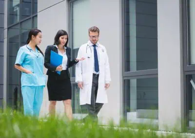 Three professionals, including two in medical attire and one in business attire holding a clipboard, walk and converse outside a modern building.