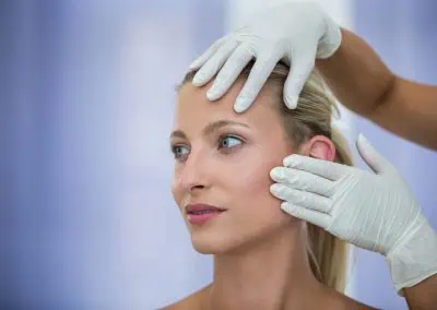 A woman's face is gently examined by gloved hands, one at her forehead and the other at her cheek.