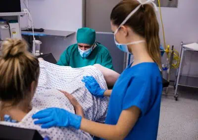 A medical team assists a woman in childbirth. The woman lies on a hospital bed with a healthcare professional in green scrubs attending to her and another in blue scrubs offering support.