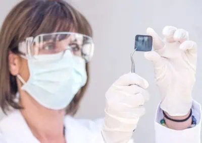 A person in lab coat, safety goggles, and a mask examines a small square object with tweezers.