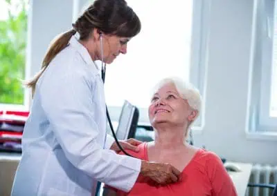 Doctor in a white coat using a stethoscope to examine an older woman in an orange shirt, while the patient sits and looks up at the doctor in a bright medical office.