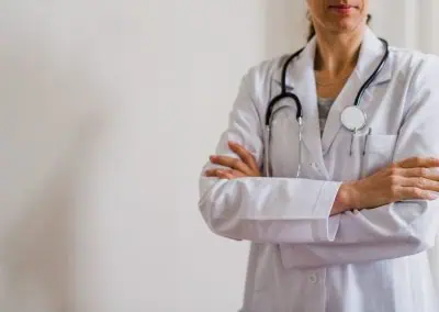 A doctor wearing a white lab coat and stethoscope stands with arms crossed against a plain background.