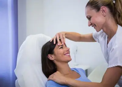 A woman in a blue shirt sitting on a chair smiling as another woman in a white uniform examines her face.