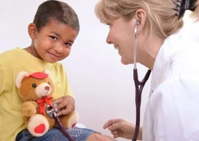 A Paediatrician in Cayman uses a stethoscope on a teddy bear held by a smiling child during a medical check-up.