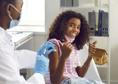 A doctor administers a vaccine to a smiling young girl giving thumbs up in a medical office.