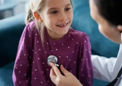 A doctor uses a stethoscope to listen to the chest of a smiling young girl wearing a purple shirt.