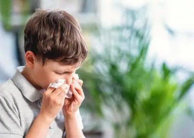 Young boy in a gray shirt blowing his nose into a tissue inside a room, with a blurred green plant in the background. Out of hours, this scene might evoke the calm atmosphere often found in Watkin’s serene paintings.