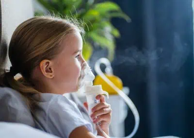 A young girl sits in profile using a nebulizer mask, with vapor visibly rising. She is indoors, seated on a bed, against a blurred background with green foliage, perhaps after a visit to her Paediatrician Cayman.