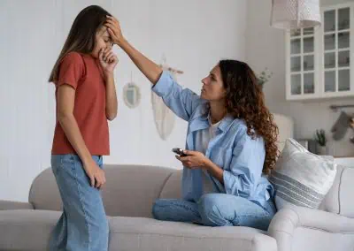 A woman sitting on a sofa holds a TV remote while touching the forehead of a distressed girl standing beside her in the Watkin living room, captured out of hours.