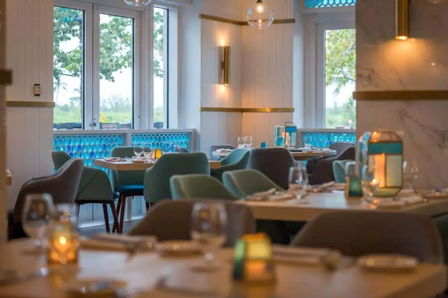 A well-lit dining area with neatly arranged tables, cushioned chairs, and decorations, featuring windows that offer an outdoor view.