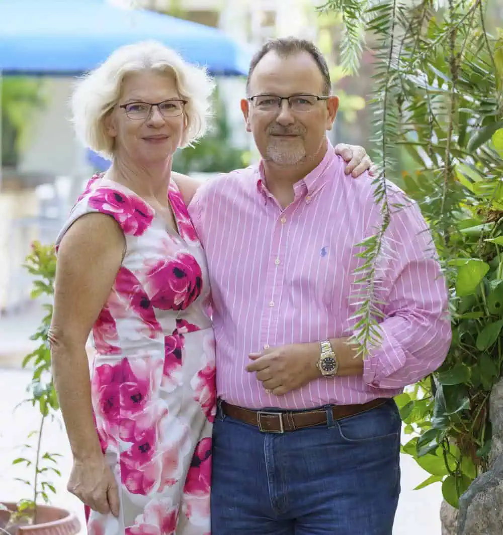 Andrew and Sara pose together outdoors. The woman is wearing a white dress with pink floral patterns, and the man is wearing a pink striped shirt and jeans. Greenery is in the background.