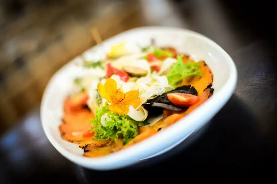 A close-up of a white bowl containing a mixed salad with leafy greens, cherry tomatoes, sliced vegetables, and an edible yellow flower garnish.