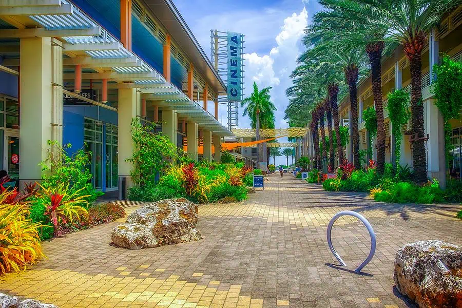 Outdoor shopping center with a cinema sign, surrounded by lush plants and palm trees. The walkway is made of patterned tiles and features scattered seating areas.