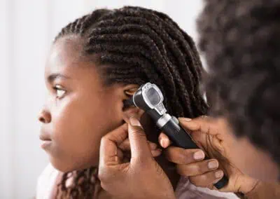 A healthcare provider is conducting a school health check in Cayman, examining a young girl's ear using an otoscope. The girl is seated, and her ear is being gently pulled back for the examination.