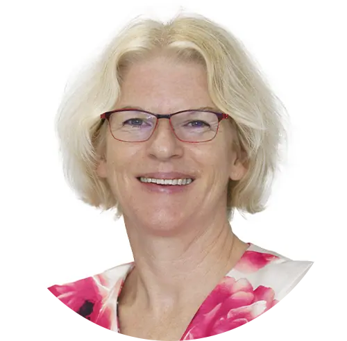 A woman with short, blond hair and glasses is smiling. She is wearing a white top with pink floral patterns. The background is plain white, suggesting a serene environment often associated with Newborn Care in Cayman.