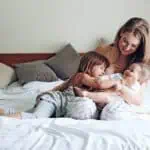 A woman sits on a bed with two young children, one standing and the other being held in her arms. They appear to be interacting and smiling.