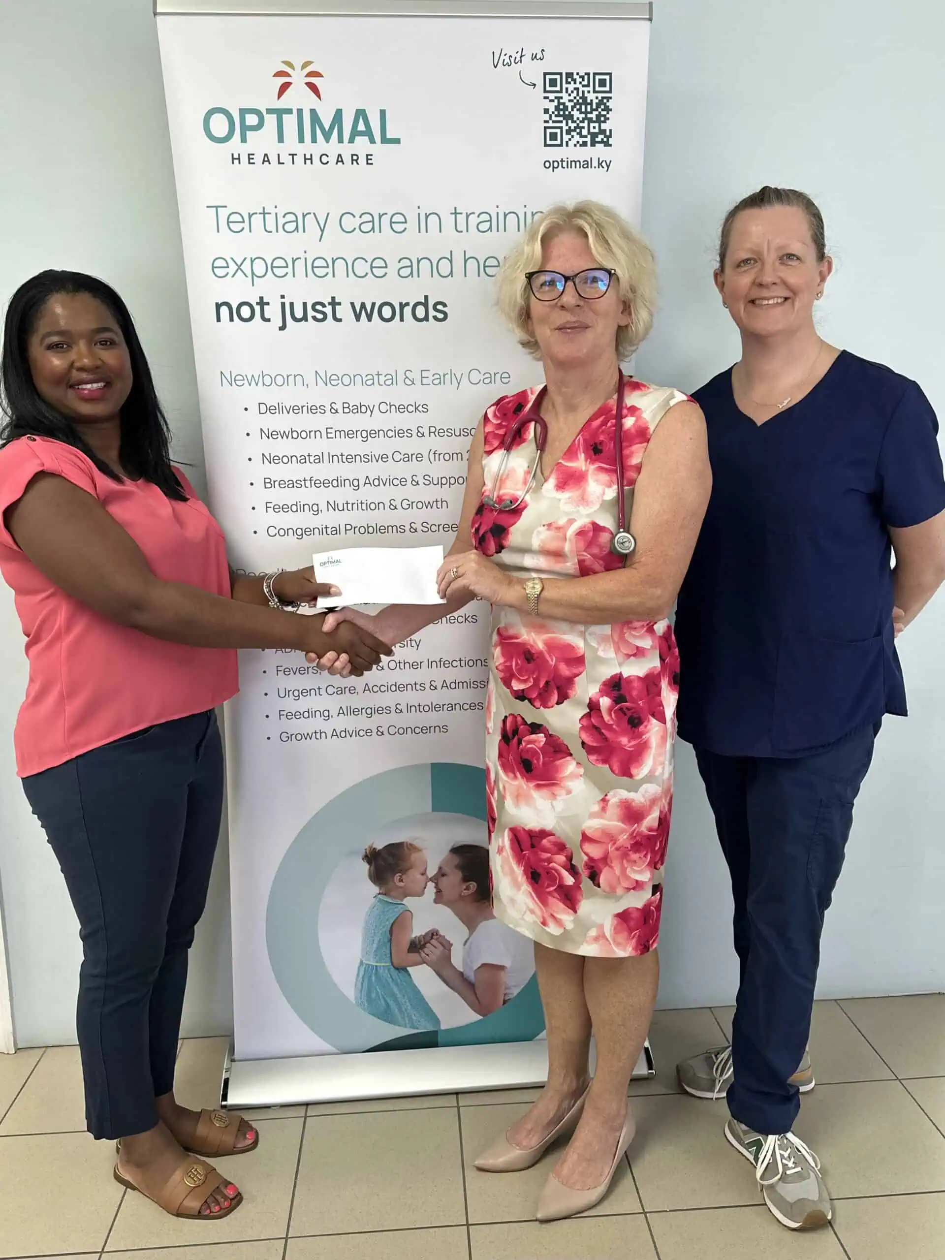 Three women standing in front of a healthcare banner, one handing a document to another. The banner advertises services for newborns, neonatal and early care.