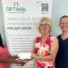 Three women standing in front of an Optimal Healthcare banner shake hands, with one giving a piece of paper to another. The banner highlights the healthcare services provided by the organization.