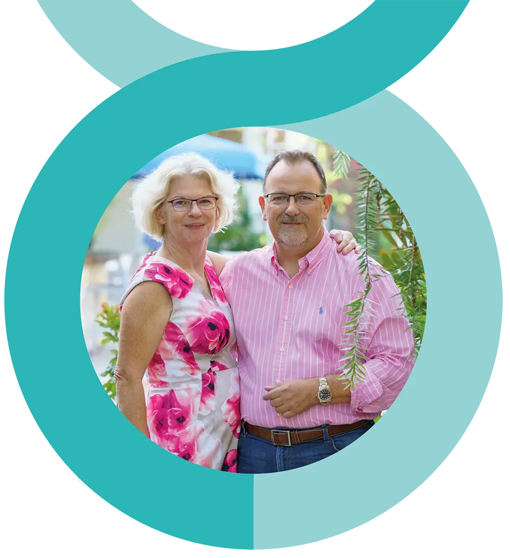 A man and woman stand closely together, smiling at the camera. The woman wears a pink floral dress, and the man wears a pink striped shirt and glasses. They are outdoors with greenery in the background.