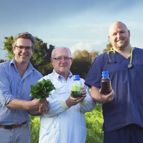 Three men standing in a field hold different items: a bunch of greens, a container of green liquid, and a container of brown liquid. They smile towards the camera.