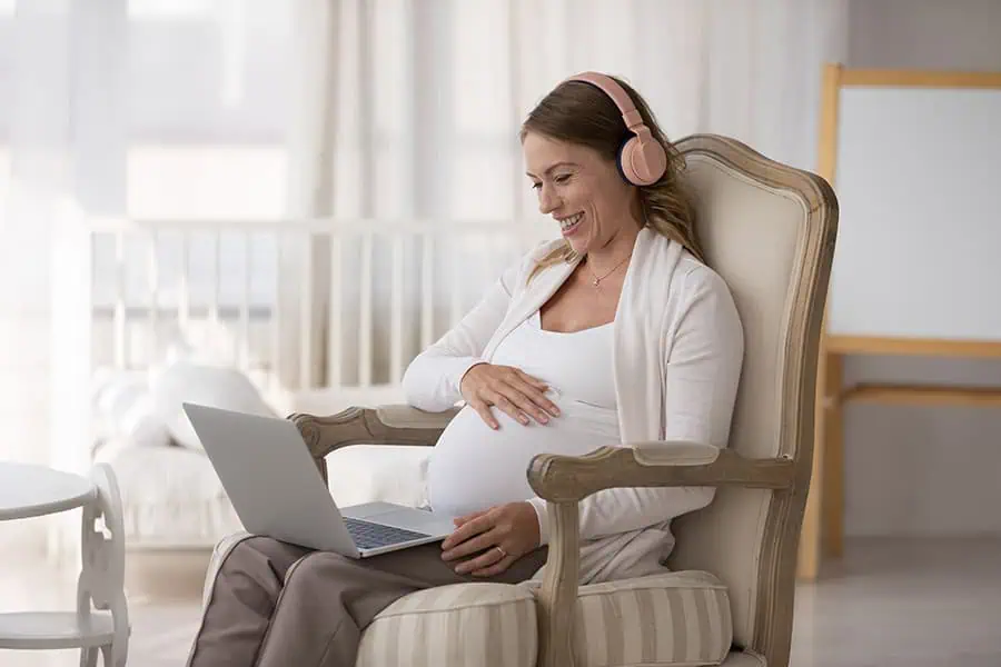 A pregnant woman sits in an armchair, wearing headphones and using a laptop. She is smiling and touching her belly. A crib is visible in the background.