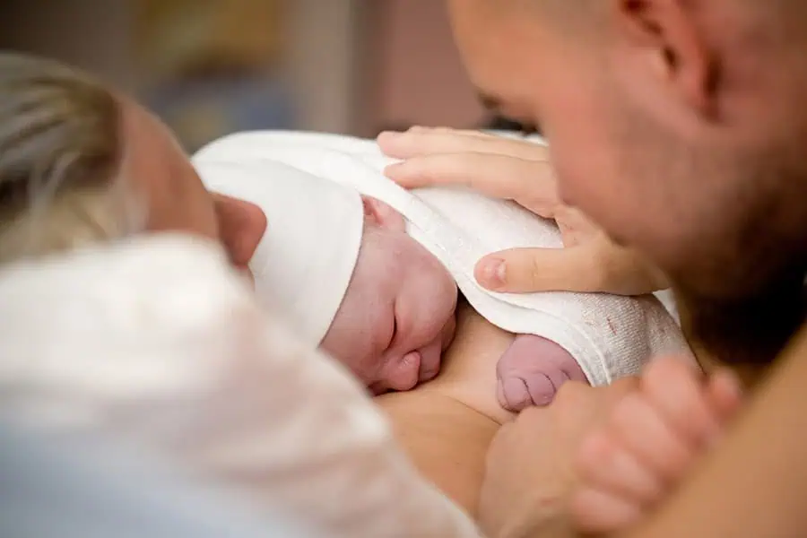 A newborn baby with a white cap sleeps on its mother's chest while a person gently touches the baby.