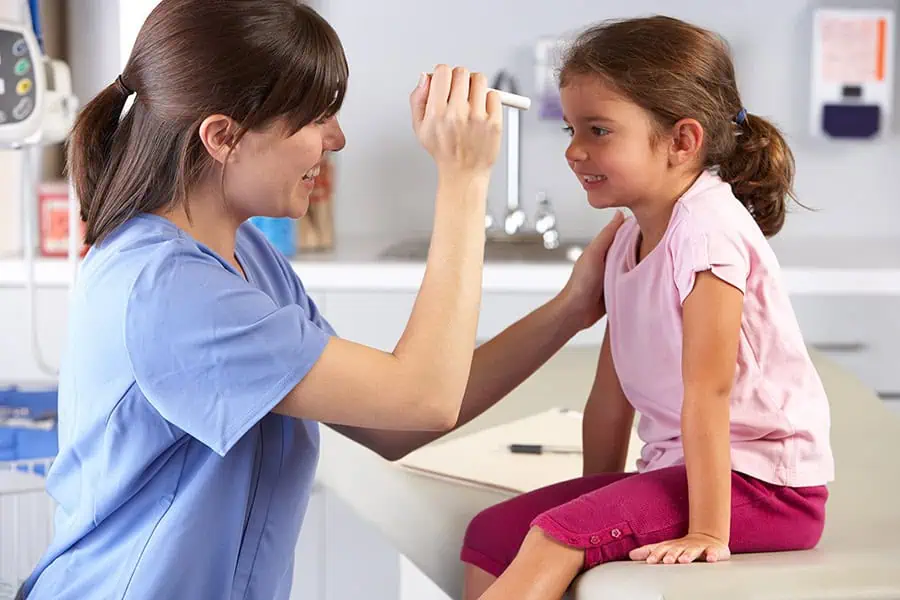 Healthcare professional performing an eye exam on a smiling young girl sitting on a medical examination table.