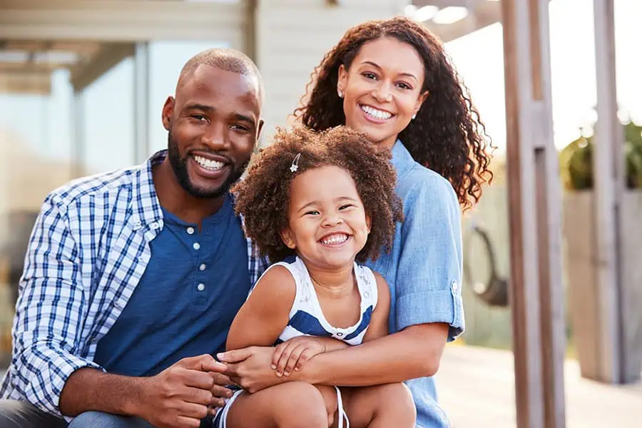 A smiling family of three, seated outside a house. The father and mother flank their child, who is in the center. Sunlight filters through, suggesting a warm, sunny day.