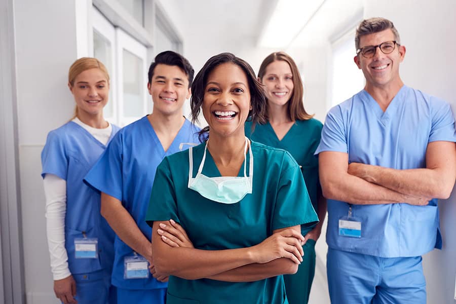 Five healthcare professionals in scrubs stand together in a hallway, with one woman smiling at the front and the others behind her, all looking at the camera.