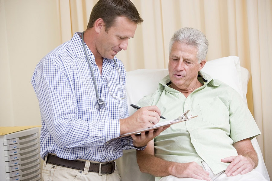 A doctor stands beside an older male patient sitting on an exam table, writing on a clipboard while they discuss medical information.