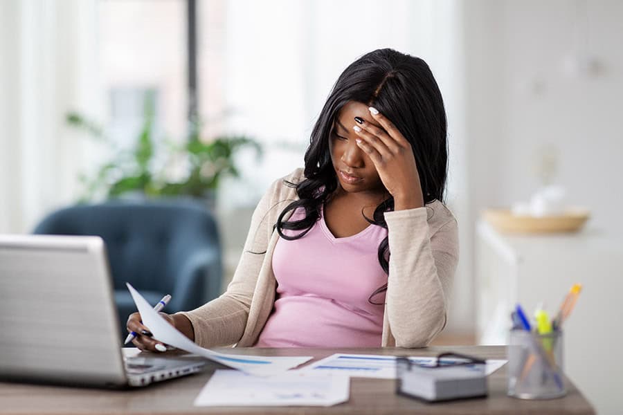A woman sits at a desk with a laptop and papers, holding her head with one hand and looking stressed or tired.